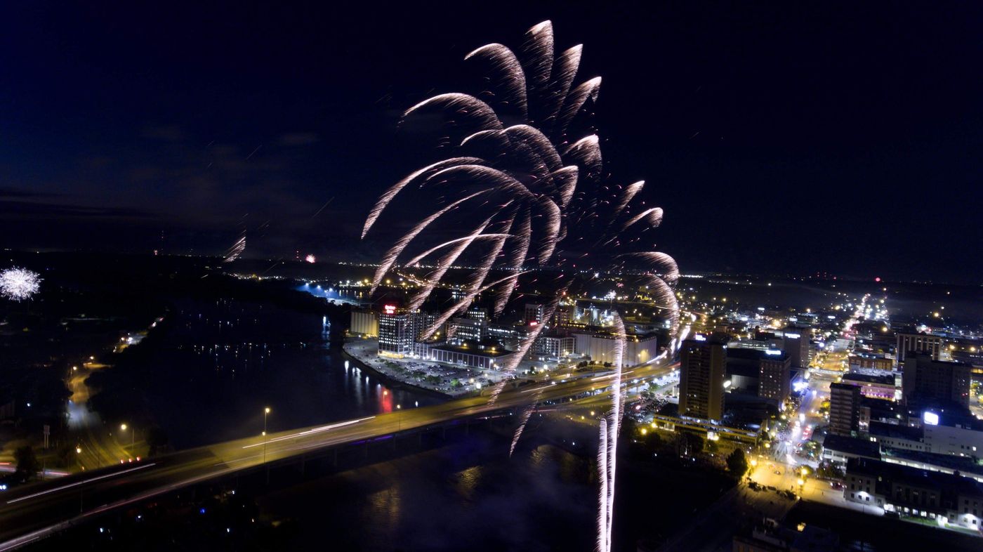 July 4 fireworks bursting over a river and downtown at night, with bridge light trails